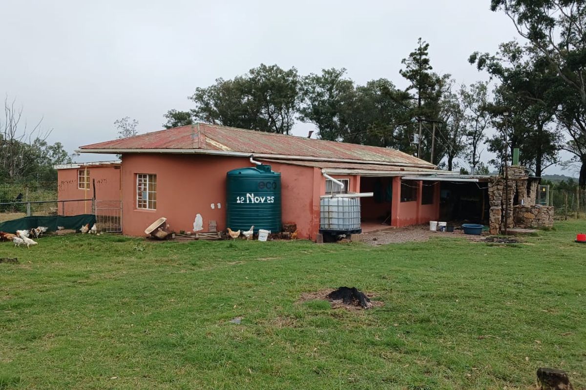 Rear view of a farm dwelling featuring a corrugated metal roof, water storage tanks, and a stone-clad outdoor fireplace set within a rural landscape.