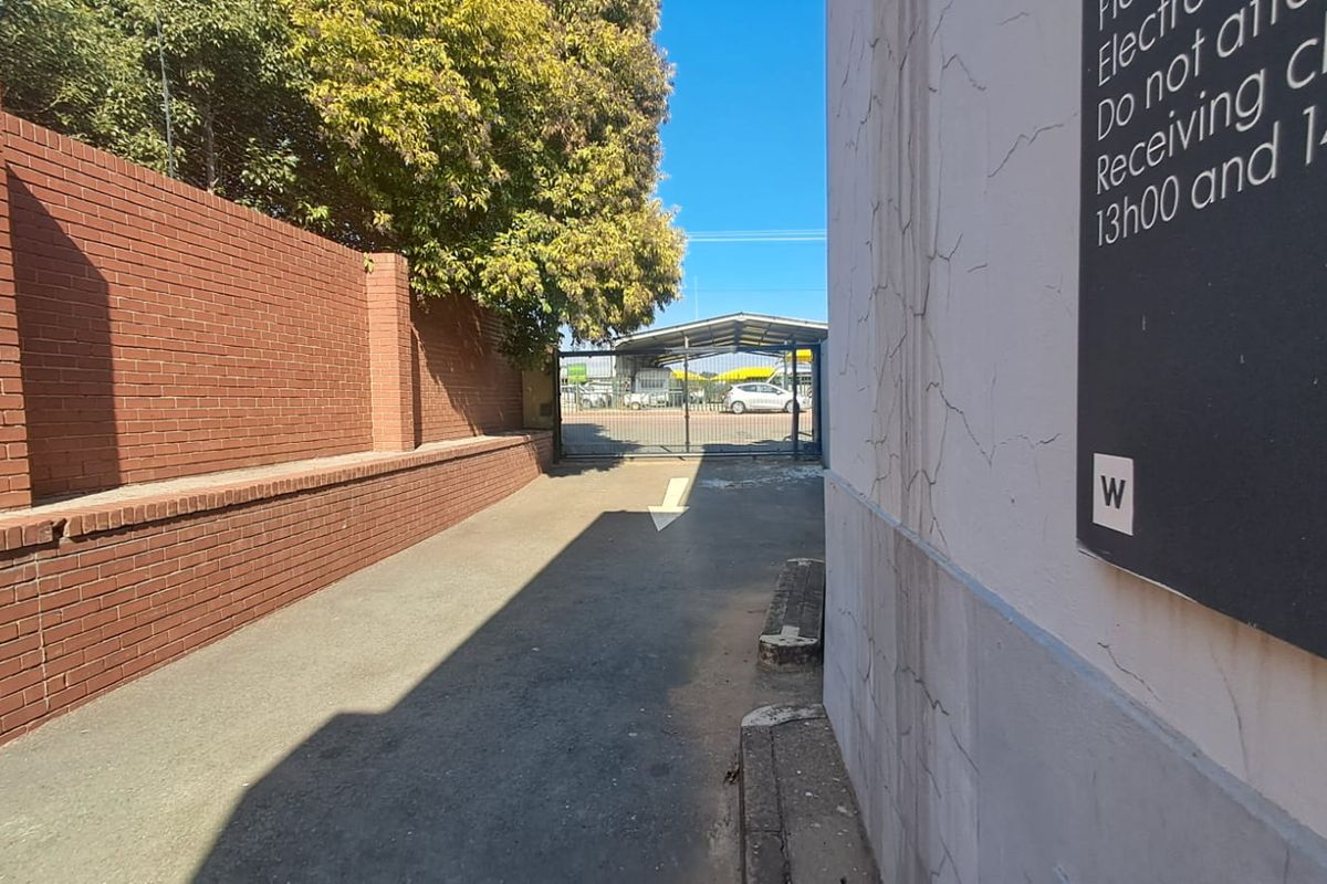 A paved service alleyway leading to a gated rear entrance, featuring a brick boundary wall and signage on the adjacent building exterior.