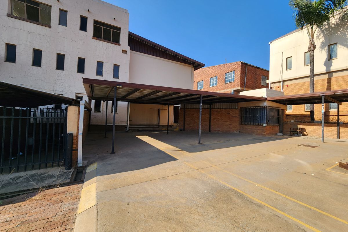 Industrial exterior area featuring covered parking bays with steel structures, concrete paving, and multi-story brick and plaster building facades.