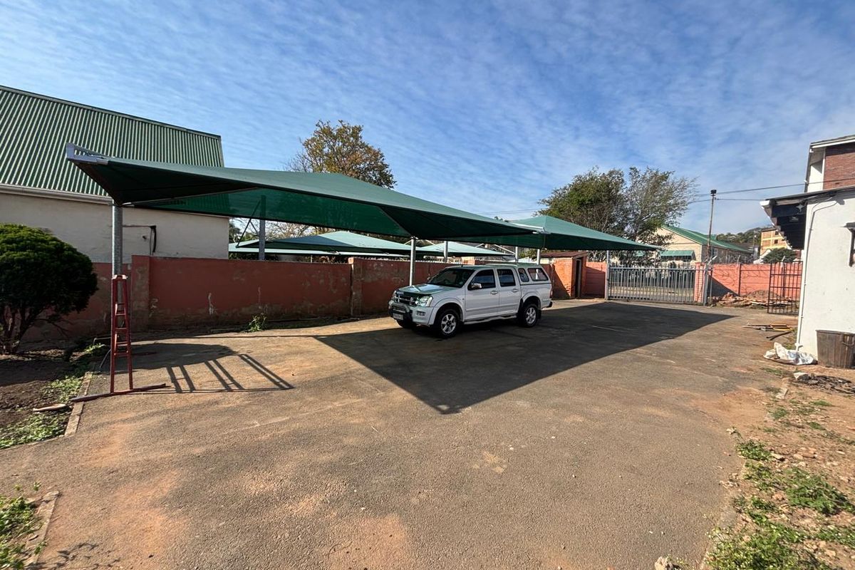 Outdoor parking area featuring a large shade cloth carport structure, asphalt paving, and perimeter boundary walls with a metal security gate.