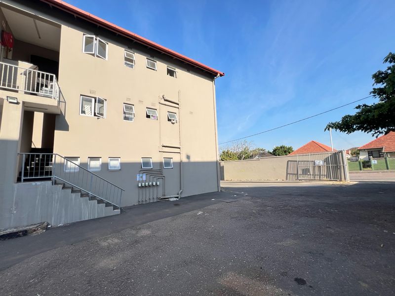Exterior view of the apartment building facade featuring a concrete staircase, metal railings, and paved parking area with perimeter walling.
