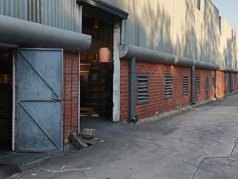 Industrial warehouse exterior featuring corrugated metal cladding, brick lower walls, ventilation louvres, and a concrete driveway with visible surface wear.
