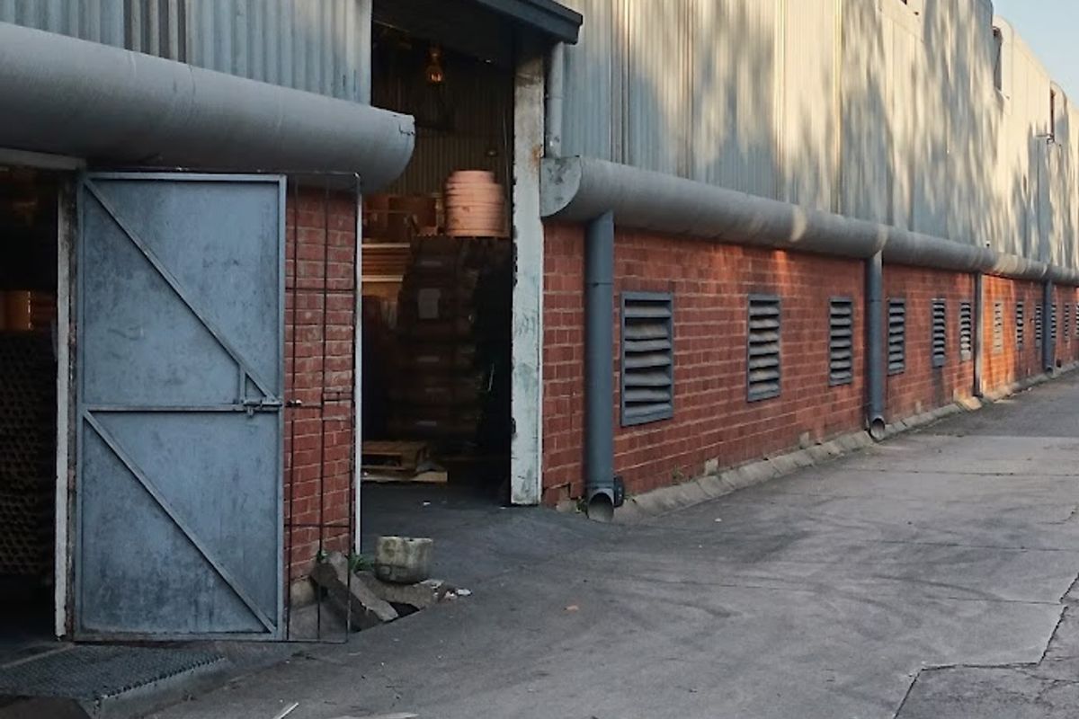 Industrial warehouse exterior featuring corrugated metal cladding, brick lower walls, ventilation louvres, and a concrete driveway with visible surface wear.
