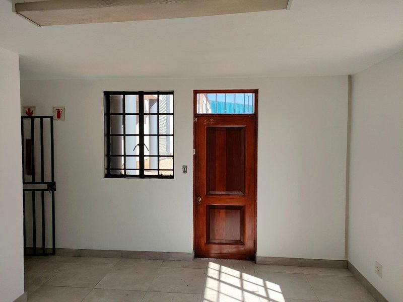 A functional industrial office space featuring tiled flooring, a secure metal-framed window, and a solid timber entrance door with a transom window.
