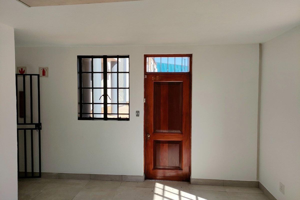 A functional industrial office space featuring tiled flooring, a secure metal-framed window, and a solid timber entrance door with a transom window.