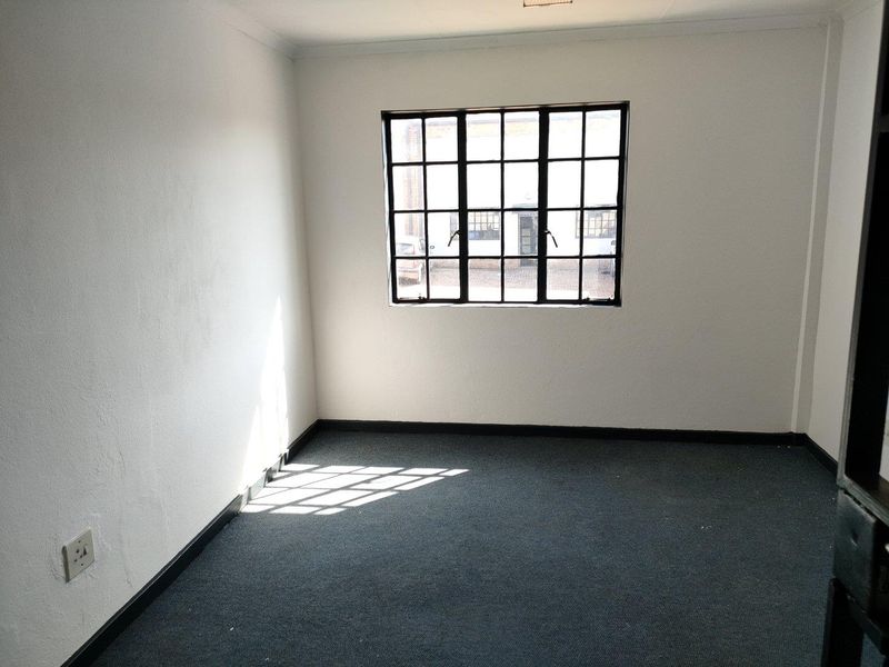 A vacant office space featuring dark carpeting, white walls, and a large industrial-style steel-framed window providing natural light.