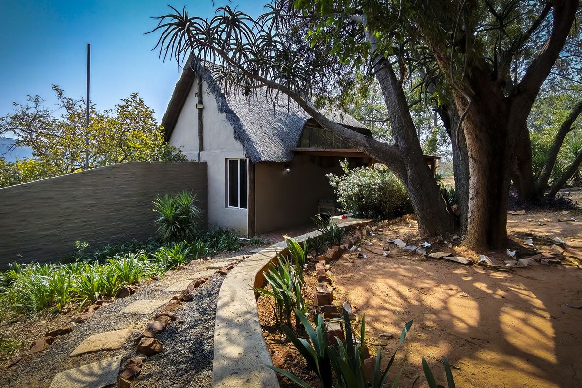 A rustic farm cottage featuring a traditional thatched roof, set within a natural landscape with a curved retaining wall and mature trees.