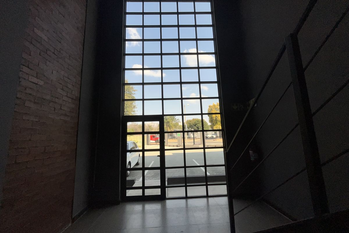 Modern office entrance featuring a double-volume grid-pattern glass window, exposed brick accent wall, and minimalist metal staircase railing.