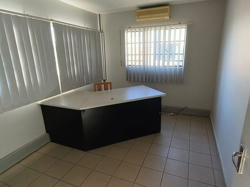 A functional office space featuring tiled flooring, vertical blinds, security bars on windows, and a wall-mounted split-system air conditioning unit.