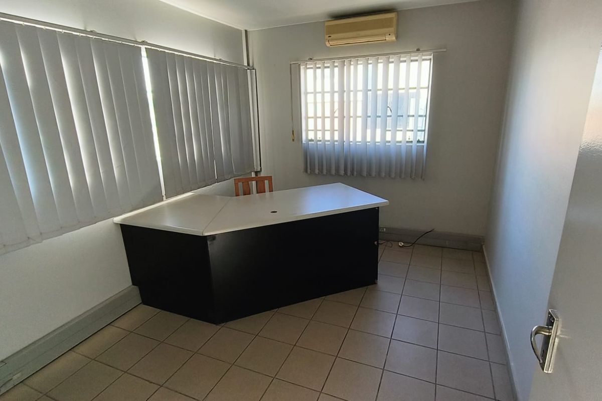 A functional office space featuring tiled flooring, vertical blinds, security bars on windows, and a wall-mounted split-system air conditioning unit.