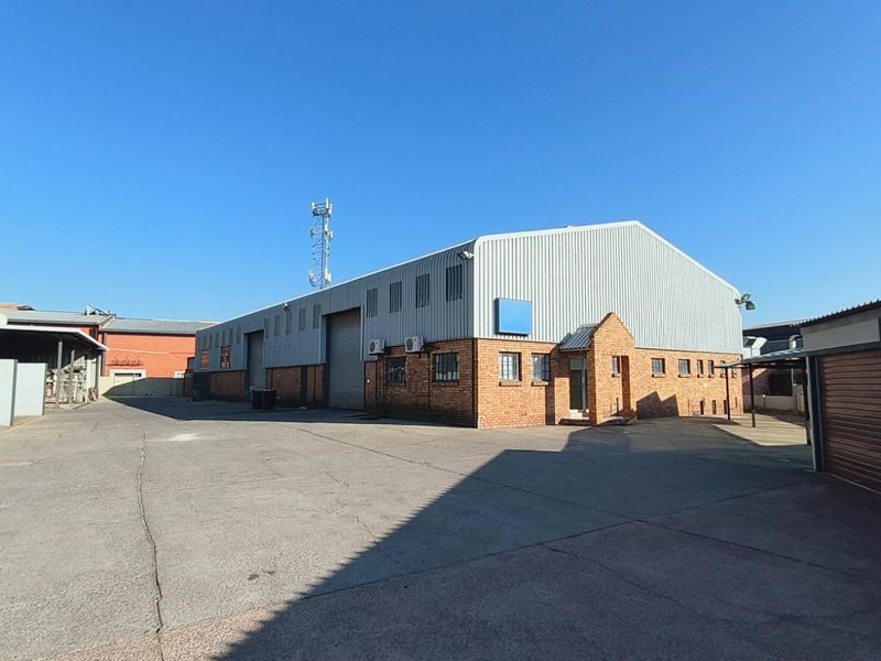 Industrial warehouse building featuring a brick facade, corrugated metal roofing, roller shutter access, and a spacious asphalt-paved yard area.