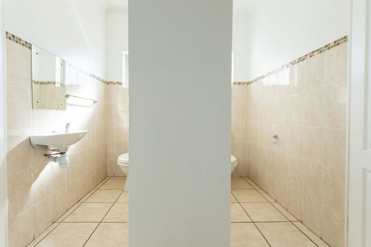 A functional office restroom featuring dual toilet stalls separated by a central partition, wall-mounted basin, and neutral ceramic tiling.