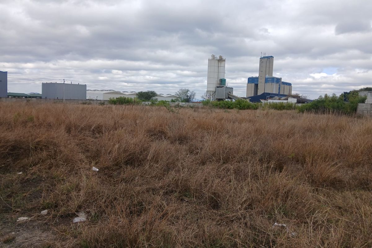 A view of a vacant commercial land plot featuring overgrown grass and perimeter fencing, with industrial structures visible in the background.