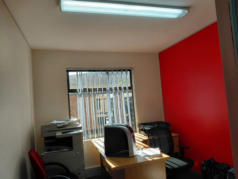 A functional office space featuring a bold red accent wall, a window with vertical blinds, and a standard fluorescent ceiling light fixture.