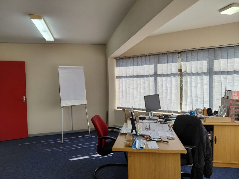 A functional office space featuring carpeted flooring, large windows with vertical blinds, and overhead fluorescent lighting.