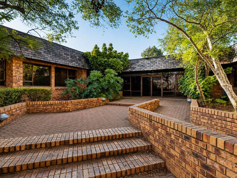 Front entrance of a brick office building featuring a paved courtyard, tiered brick retaining walls, and large glass doors under a slate roof.