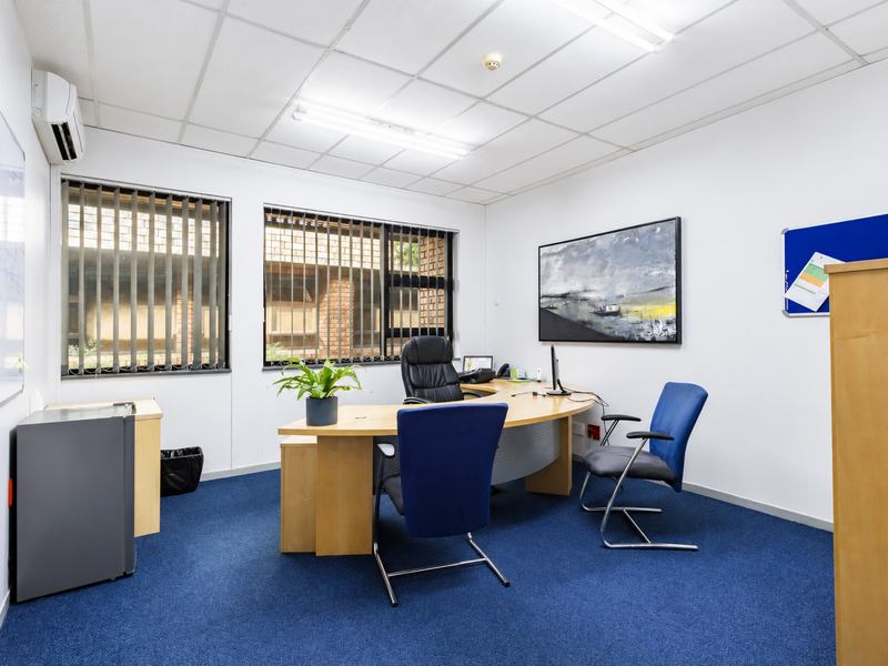 A functional office space featuring a suspended ceiling, fluorescent lighting, wall-mounted air conditioning, and large windows with vertical blinds.
