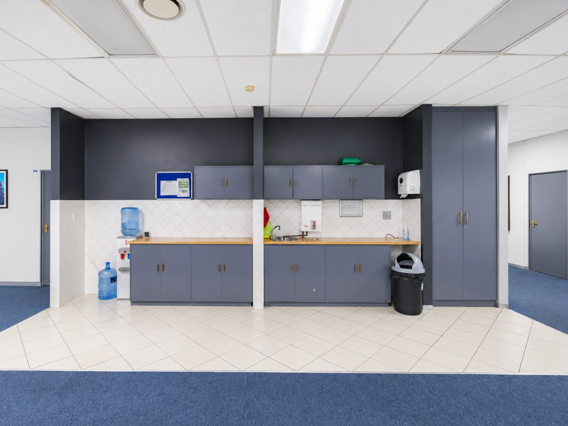 A functional office kitchenette featuring grey built-in cabinetry, laminate countertops, a stainless steel sink, and a tiled backsplash with a suspended ceiling.
