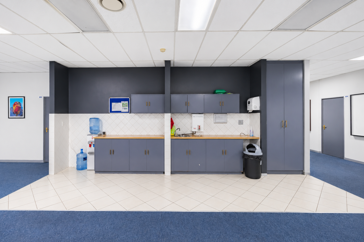 A functional office kitchenette featuring grey built-in cabinetry, laminate countertops, a stainless steel sink, and a tiled backsplash with a suspended ceiling.
