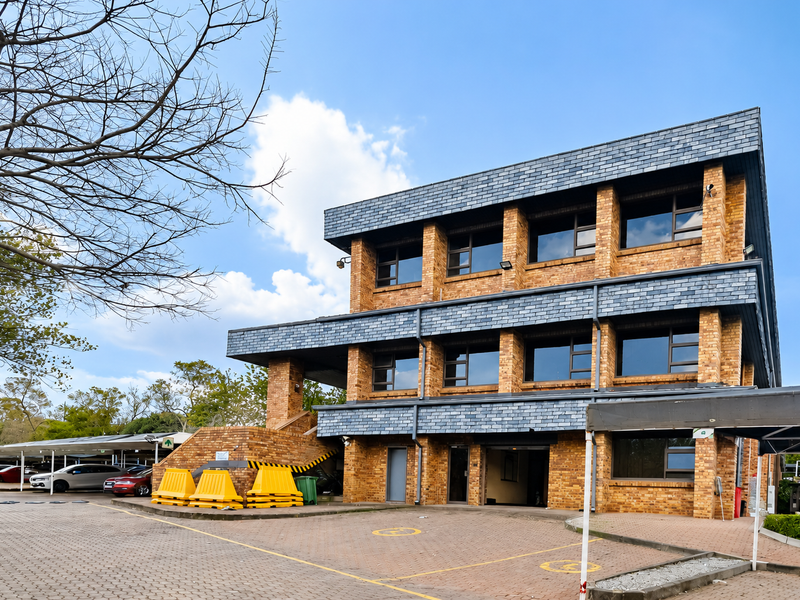 A three-story office building featuring a face brick facade, slate roof detailing, and an adjacent paved parking area with covered bays.