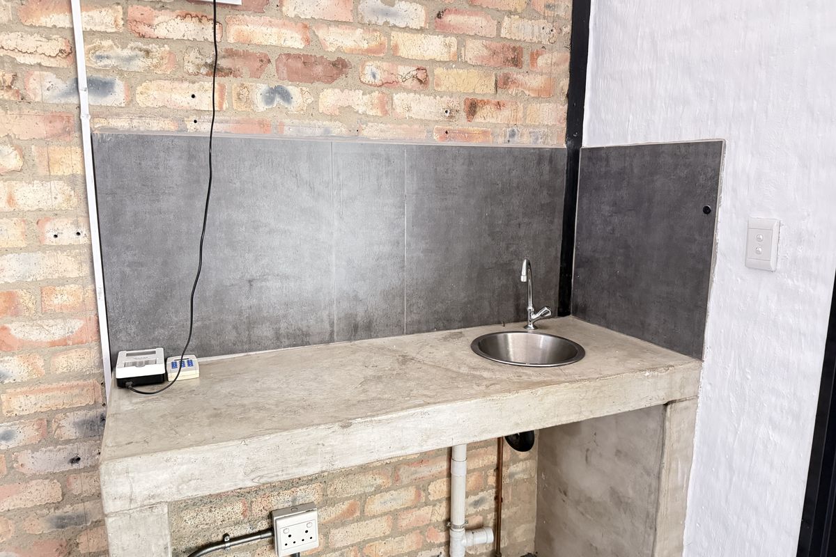 A functional kitchenette area featuring a concrete countertop, integrated stainless steel sink, and dark grey splashback tiles set against an industrial brick wall.