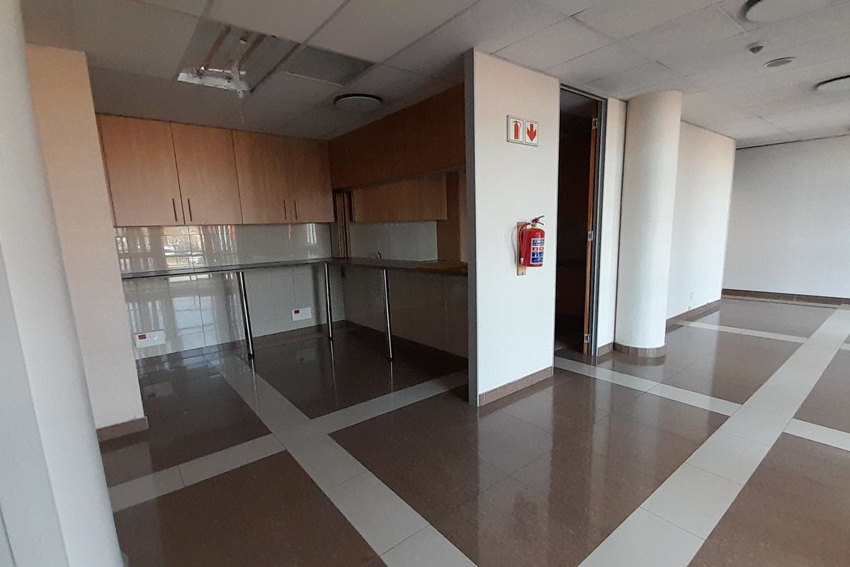 A functional office kitchenette area featuring built-in cabinetry, tiled flooring, and a suspended ceiling, currently vacant and ready for use.