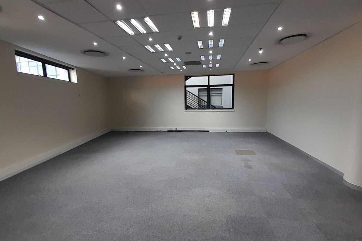 A spacious, empty office area featuring a suspended ceiling with recessed lighting, carpeted flooring, and power skirting along the walls.