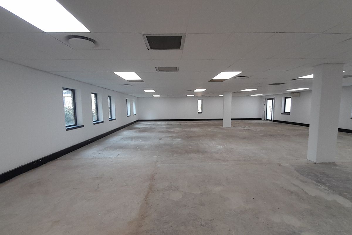 A large, open-plan office space featuring a suspended ceiling with recessed lighting, structural columns, and unfinished concrete flooring.