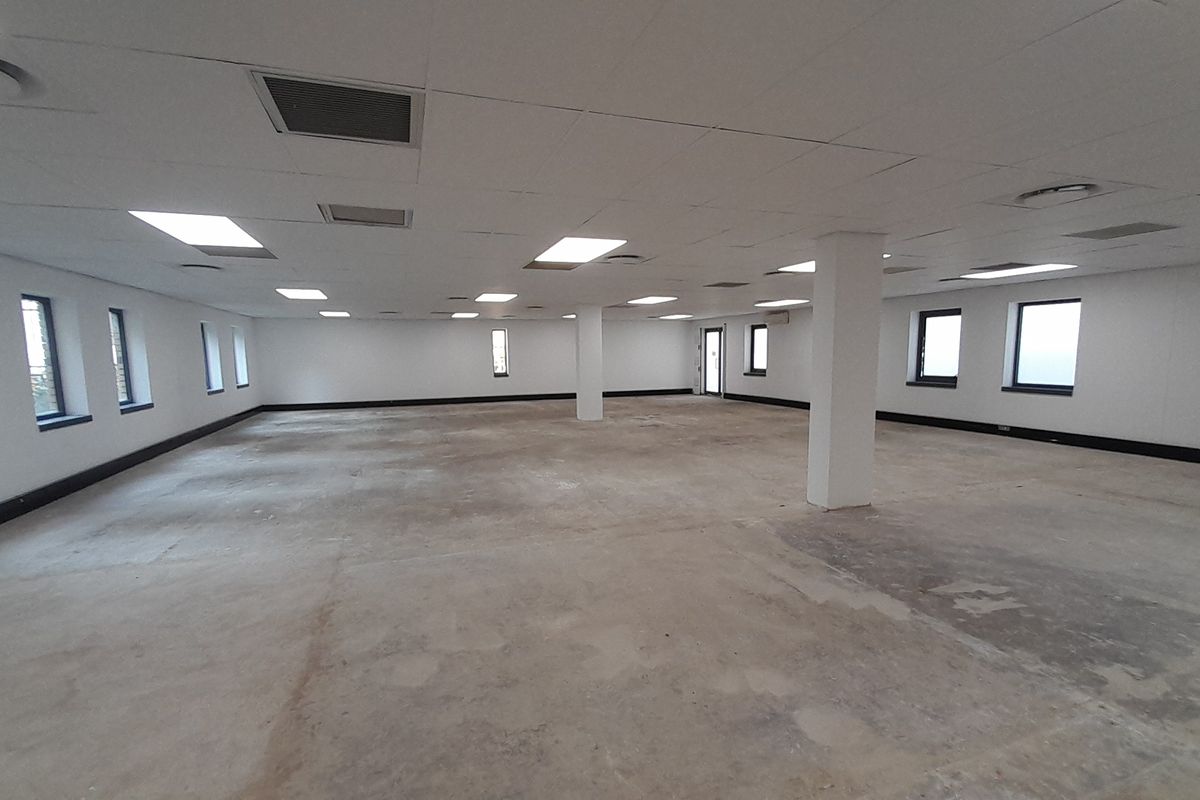A large, open-plan office space featuring a suspended ceiling, recessed lighting, structural columns, and expansive windows, currently awaiting flooring installation.