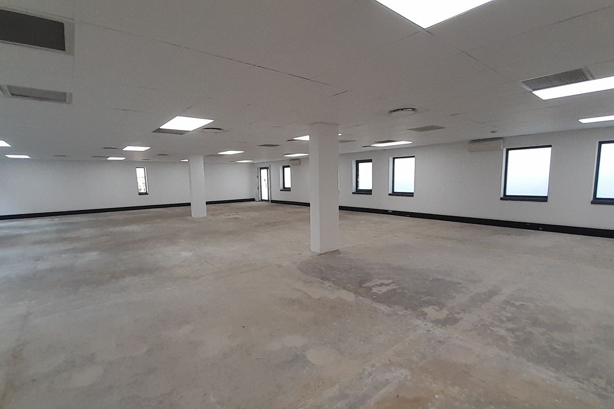 A large, open-plan office space featuring a suspended ceiling, recessed lighting, and structural columns, currently vacant with unfinished concrete flooring.
