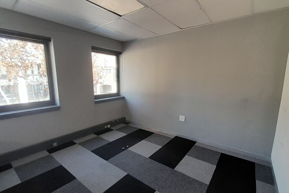 An empty office space featuring a suspended ceiling, grey and black carpet tiles, and natural light from two windows.