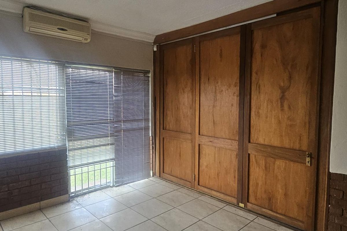 A bedroom featuring built-in wooden cupboards, tiled flooring, and a wall-mounted air conditioning unit with large windows.