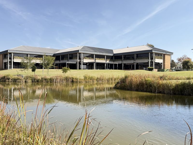 A wide-angle view of a modern industrial office building featuring a metal roof, concrete structural pillars, and extensive glass frontage overlooking a landscaped pond.