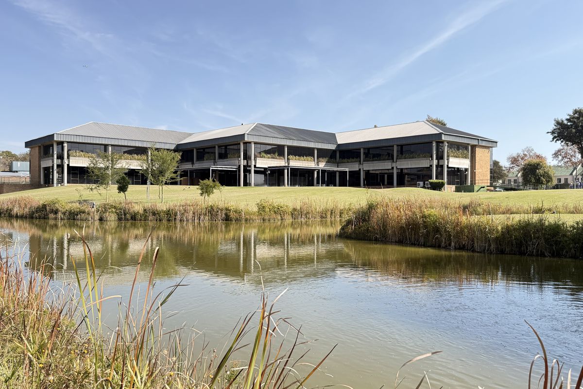 A wide-angle view of a modern industrial office building featuring a metal roof, concrete structural pillars, and extensive glass frontage overlooking a landscaped pond.