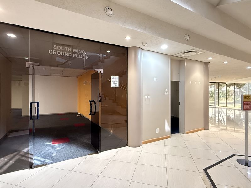 Spacious commercial building entrance lobby featuring tiled flooring, recessed lighting, and a glass-partitioned access point to the South Wing ground floor.