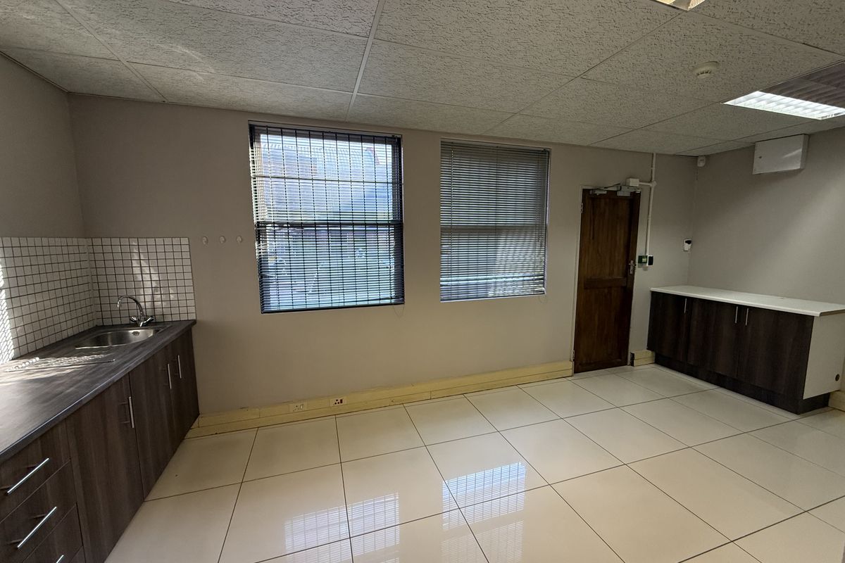 A functional commercial kitchenette featuring tiled flooring, built-in cabinetry with laminate countertops, a stainless steel sink, and a drop ceiling with integrated lighting.