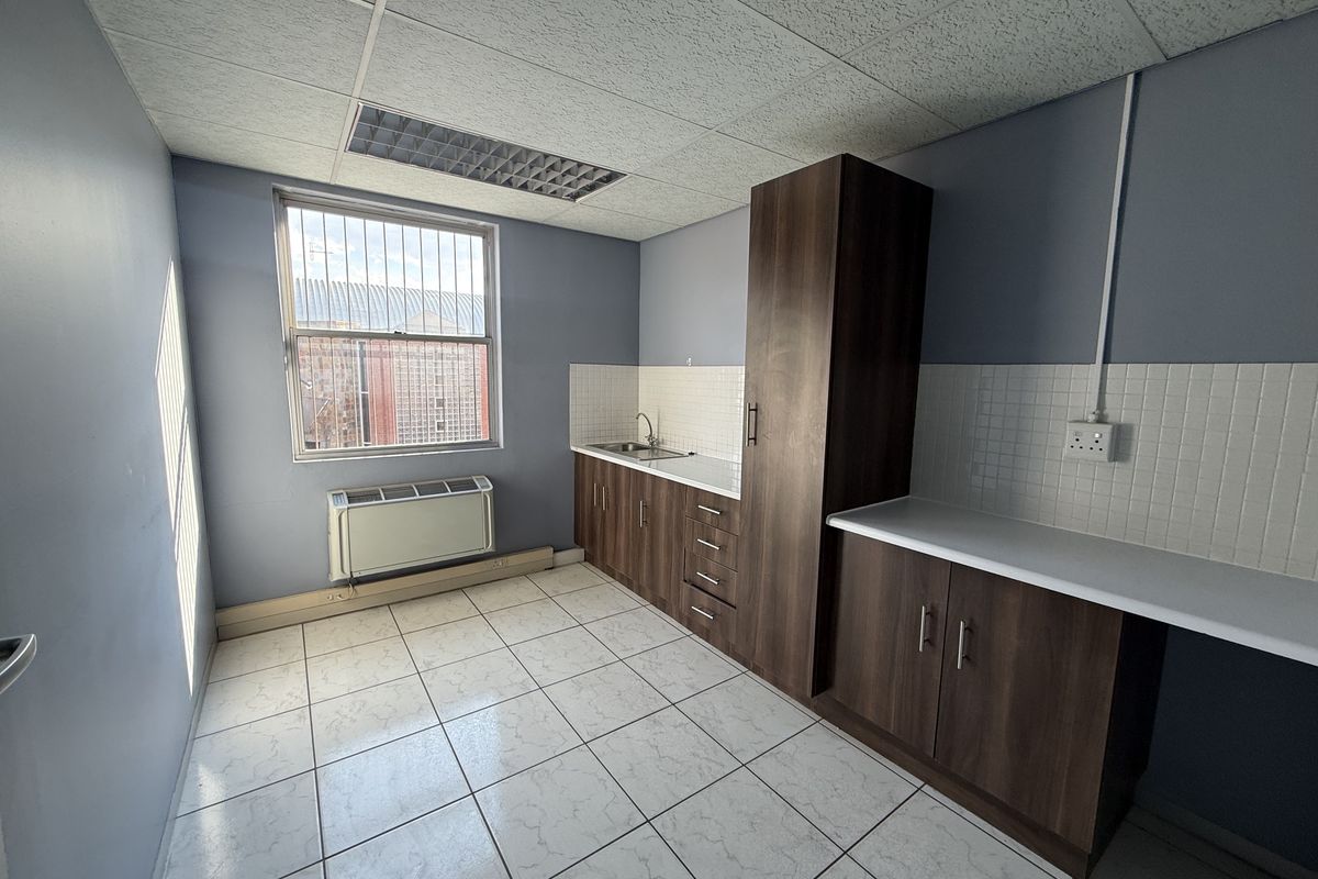 A functional commercial kitchenette featuring dark wood cabinetry, laminate countertops, a stainless steel sink, tiled backsplash, and a wall-mounted HVAC unit.