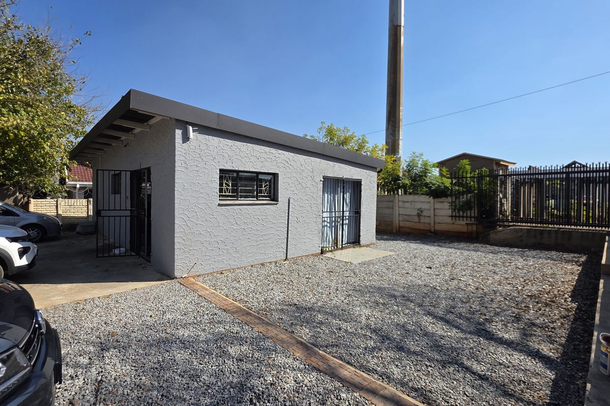 Exterior view of a grey textured office building with security gates and bars, featuring a gravel parking area and perimeter fencing.