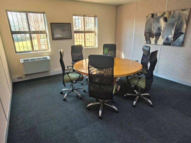 A functional office meeting room featuring carpeted flooring, wall-mounted HVAC unit, and windows with security bars.