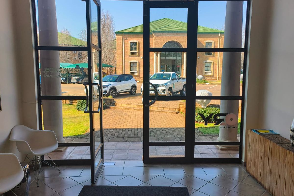 Bright office entrance featuring large glass doors, tiled flooring, and structural columns, providing a clear view of the exterior parking area.