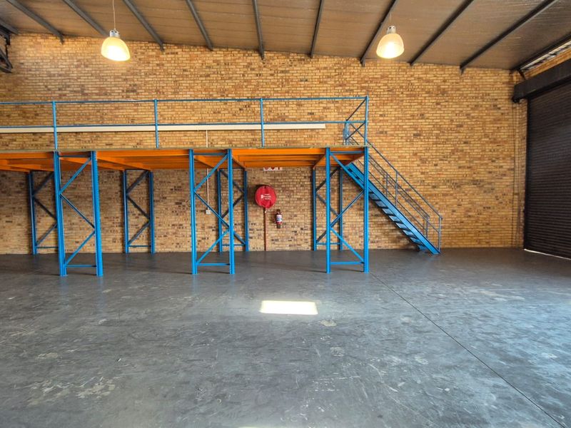 Spacious industrial warehouse unit featuring a mezzanine level, steel staircase, exposed brick walls, concrete flooring, and a large roller shutter door.
