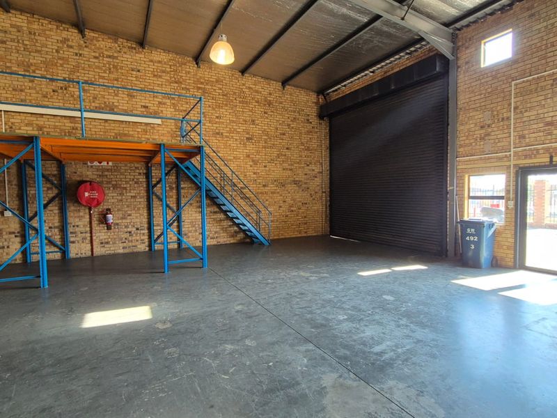 Spacious industrial warehouse unit featuring exposed brick walls, concrete flooring, a steel mezzanine level with staircase, and a large roller shutter door.