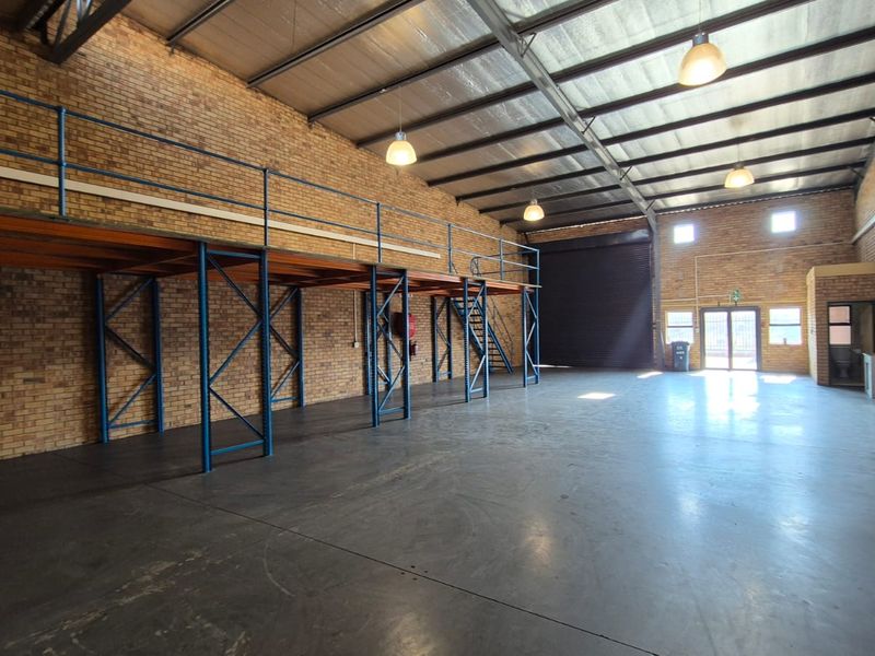 Spacious industrial warehouse unit featuring high ceilings, exposed brick walls, a functional steel mezzanine level, and a large roller shutter door for easy access.