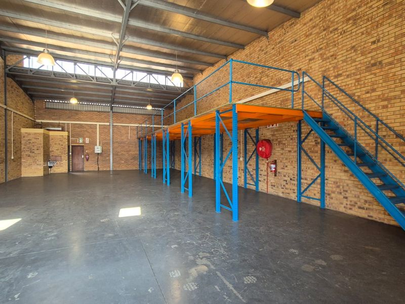 Spacious industrial warehouse unit featuring exposed brick walls, high ceilings with steel trusses, concrete flooring, and a functional mezzanine level with staircase access.