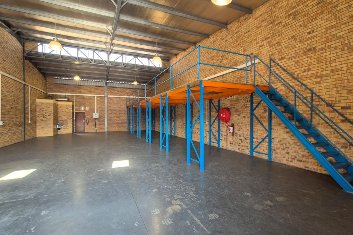 Spacious industrial warehouse unit featuring exposed brick walls, high ceilings with steel trusses, concrete flooring, and a functional mezzanine level with staircase access.