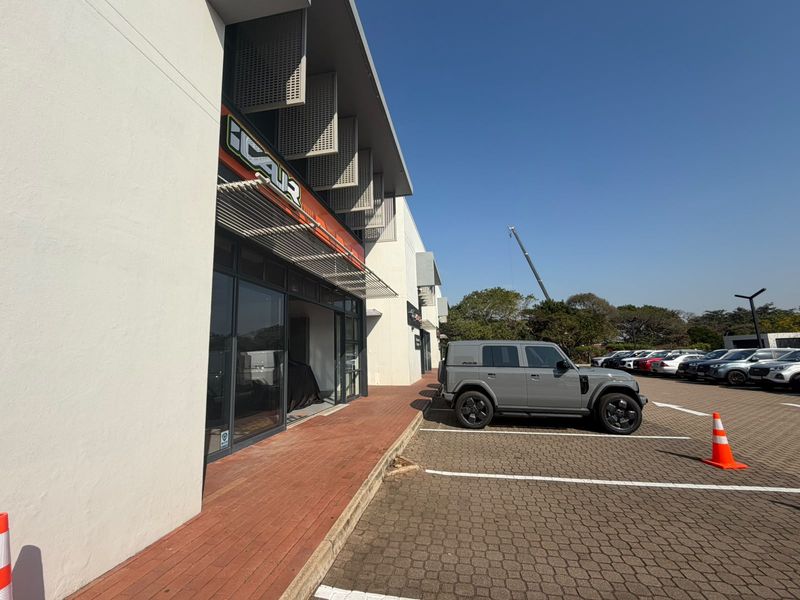 Exterior view of a retail storefront featuring a glass facade, metal louvered shades, and a paved parking area.