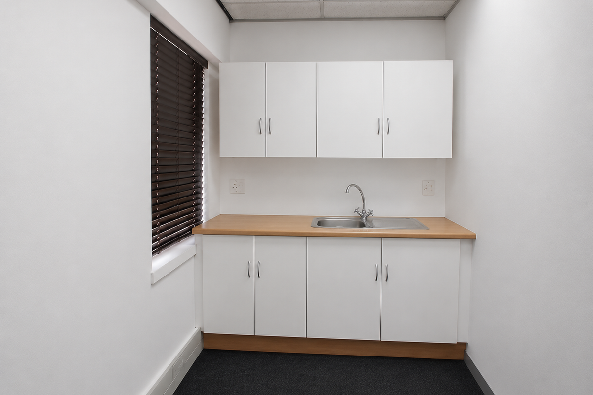 A clean, compact office kitchenette featuring white built-in cabinetry, a stainless steel sink, and a wood-look countertop.
