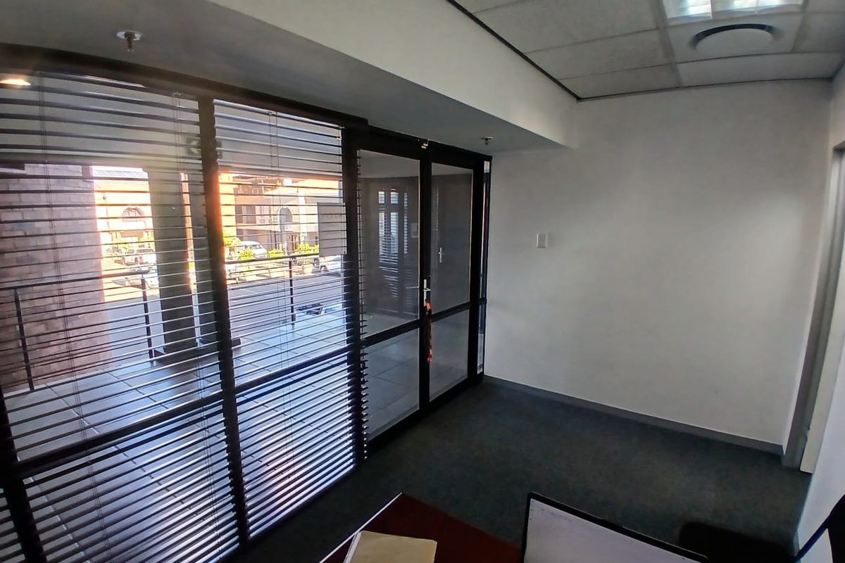 A bright, empty office space featuring carpeted flooring, a suspended ceiling with recessed lighting, and large aluminium-framed glass doors with venetian blinds.