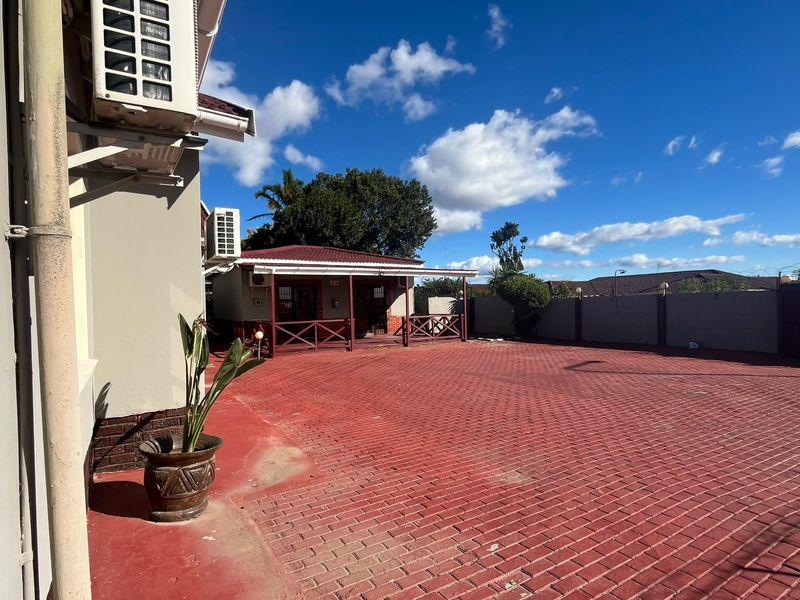A spacious rear exterior area featuring a large red paved driveway and a covered patio structure attached to the guest house building.