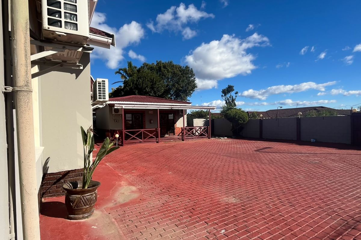 A spacious rear exterior area featuring a large red paved driveway and a covered patio structure attached to the guest house building.